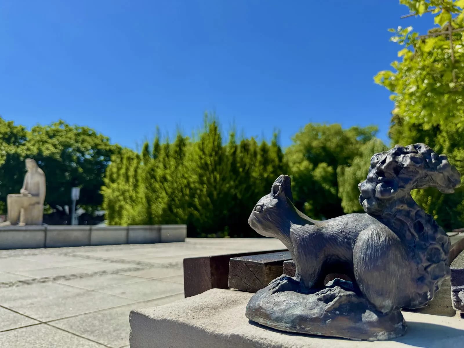 A bronze sculpture of a squirrel is in the foreground, with a large stone sculpture of a person and green trees visible in the background under a clear blue sky.
